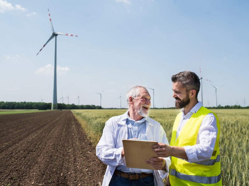 Two engineers or technicians with clipboard standing on wind farm, making notes.